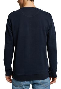 Homme portant un sweat-shirt uni bleu marine foncé à manches longues et un jean bleu clair, vu de dos sur fond blanc.