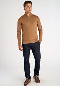 Hellbrauner Reißverschluss-Pullover mit langen Ärmeln, über marineblauen Hosen getragen. Braune Schnürschuhe mit einer strukturierten Sohle.
