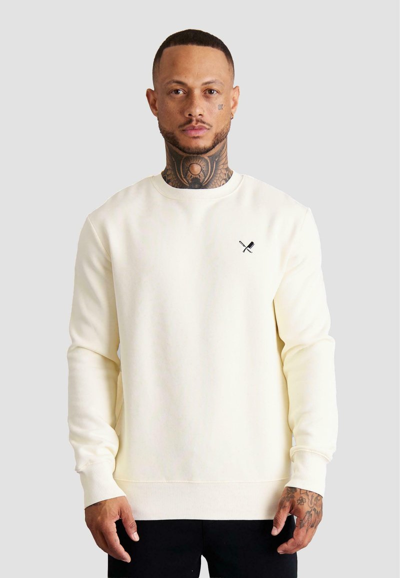 Sudadera cremosa fabricada con tejido suave, que presenta mangas largas, un cuello redondo y un pequeño logo de cuchillos cruzados en negro en el pecho.