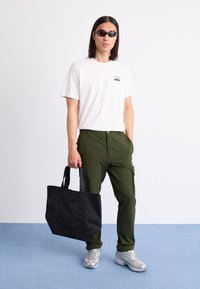 White t-shirt with a logo, green cargo pants, black tote bag, and silver sneakers. Model wears dark sunglasses, standing on a blue floor.
