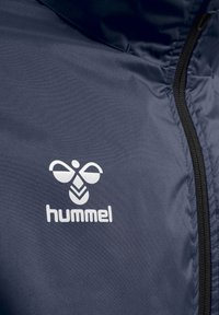 Gros plan d’une veste bleu marine avec une fermeture éclair noire et un logo blanc Hummel avec une abeille stylisée au-dessus du nom de la marque sur le tissu.