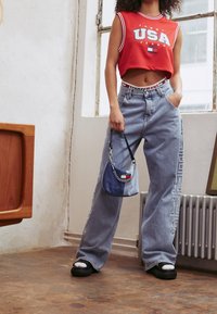 Top cortito sin mangas rojo con estampado de "USA", jeans de pierna ancha de color azul claro con detalle de logo, y un pequeño bolso cruzado azul con cadena.