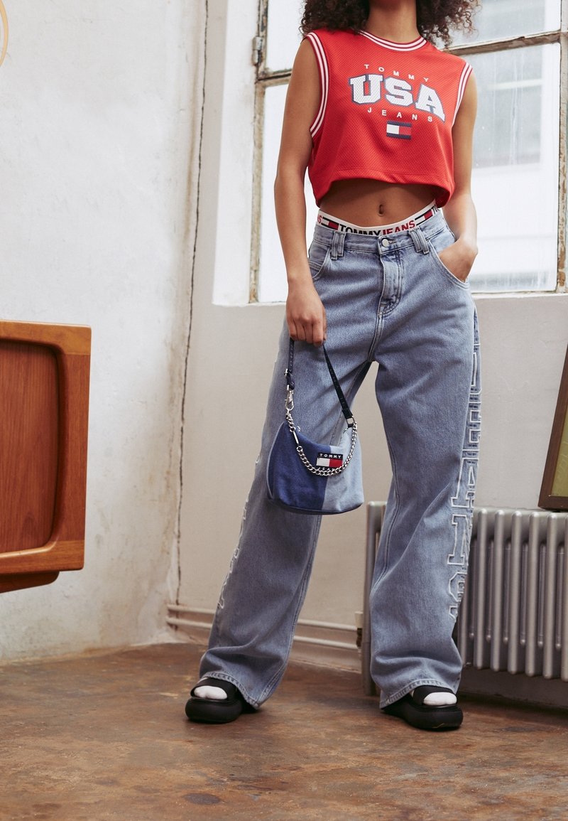 Top cortito sin mangas rojo con estampado de "USA", jeans de pierna ancha de color azul claro con detalle de logo, y un pequeño bolso cruzado azul con cadena.