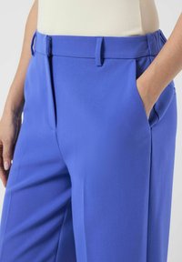 Blå bukser med skreddersydd passform, glatt stoff, frontlommer og en flat linning med beltehemper. Nærbilde av plagget vist.