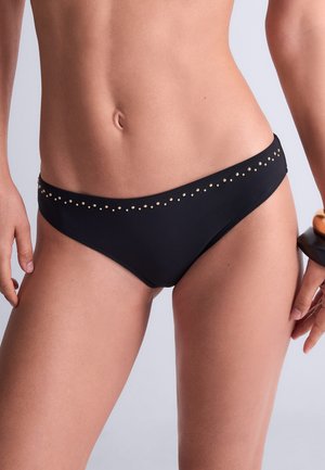 Femme portant un bas de bikini noir avec de petits clous dorés le long de la taille, visible de la taille jusqu'en haut des cuisses sur un fond uni.