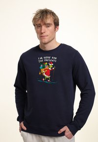 Sudadera de color navy con un gráfico de un personaje verde con un gorro de Santa esquiando, con el texto que dice "Estoy aquí por los regalos." Material de mezcla de algodón.