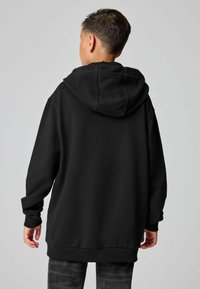 Chico con el pelo corto que lleva una sudadera negra holgada y unos jeans gris oscuro, de pie de espaldas frente a un fondo liso de color gris claro.