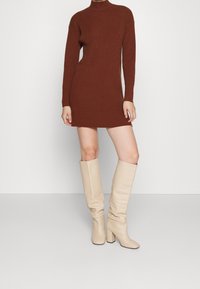 Braun geripptes Strickkleid mit langen Ärmeln und hohem Ausschnitt, kombiniert mit beigen knielangen Blockabsatzstiefeln. Glatte Textur, taillierter Schnitt.