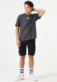 Graues kurzärmliges T-Shirt mit einem kleinen Logo, schwarze Cargo-Shorts, weiße High-Top-Sneaker und gemusterte schwarz-weiße Socken.
