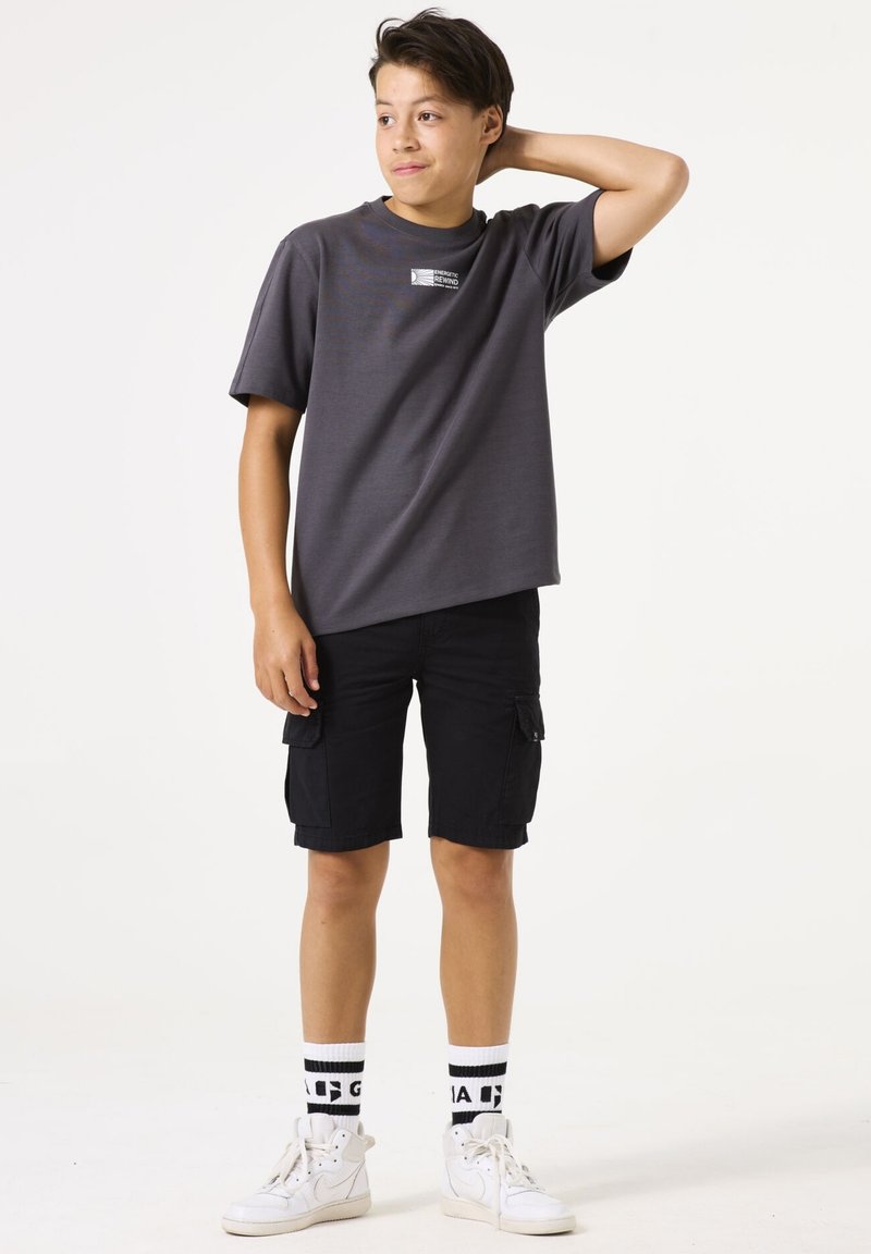 Graues kurzärmliges T-Shirt mit einem kleinen Logo, schwarze Cargo-Shorts, weiße High-Top-Sneaker und gemusterte schwarz-weiße Socken.