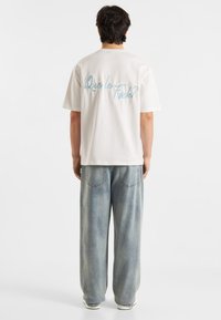Witte katoenen t-shirt met blauwe geborduurde tekst "Que le f***?" op de achterkant, gecombineerd met losse lichtblauwe jeans en witte sneakers.
