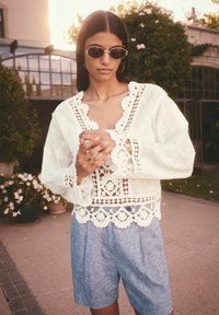 Hvid blondebluse med blomstertryk og bølgede kanter, parret med lyseblå plisserede shorts. Model holder en kop, iført solbriller.