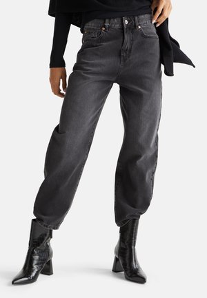 Frau trägt hochgeschnittene, lockere schwarze Jeans und schwarze spitze Stiefel mit Absatz vor weißem Hintergrund.