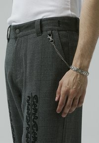 Homme portant un pantalon à carreaux gris avec imprimé de texte noir, une chaîne argentée attachée à un passant de ceinture et un bracelet en argent à motif floral au poignet droit.