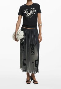 Černé tričko s motivem srdce, kombinované s plisovanou černo-šedou sukní s abstraktními vzory, drží bílou tašku a má na sobě sandály na blokovém podpatku.