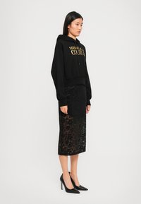Svart cropped hettegenser med gulltekst, kombinert med et svart midi skjørt i blonder med blomster mønster og gjennomsiktig bunn. Sorte spisse hæler fullfører antrekket.