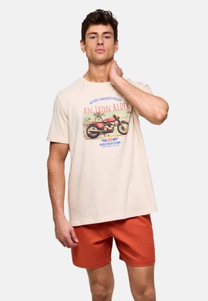 Hombre con camiseta beige con gráfico de motocicleta vintage y texto rojo, combinado con pantalones cortos naranjas, posando con la mano en el cuello.