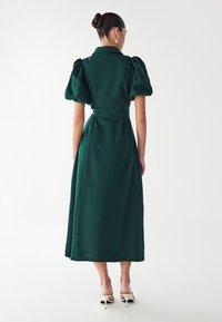 Zelena obleka do kolen, izdelana iz strukturirane tkanine, z napihnjenimi rokavi in beljenim pasom, ki ima srajčni ovratnik in raven kroj.