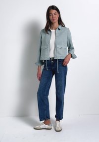Mujer con chaqueta corta azul claro, top blanco, vaqueros azules y deportivas beige y blancas, de pie frente a un fondo blanco liso.