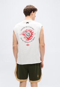 Λευκό αμάνικο μπλουζάκι με γραφικό σχέδιο, που απεικονίζει ένα πολύχρωμο λιοντάρι με κείμενο στο πίσω μέρος. Χακί σορτς με χρυσές λεπτομέρειες και λογότυπο.