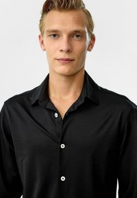 Zwarte button-up shirt met een gladde textuur, voorzien van een kraag en witte knopen. Het ontwerp is strak, met een getailleerde silhoutte die eenvoud benadrukt.