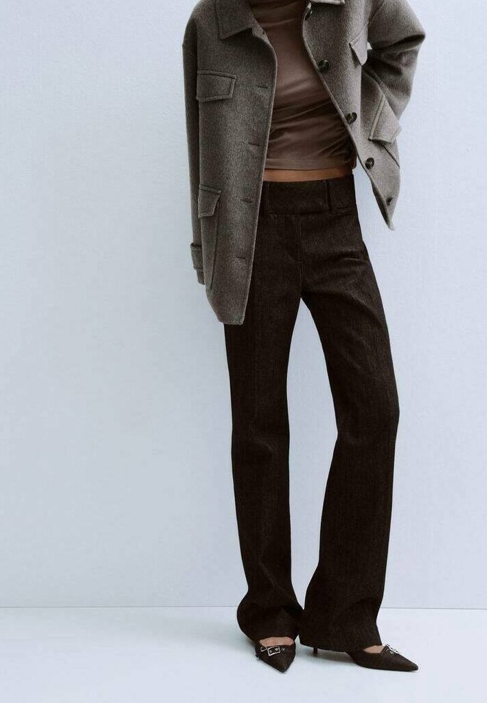 Personne portant un pantalon marron foncé, un col roulé taupe, une veste en laine grise et des talons pointus noirs, debout contre un mur gris clair.