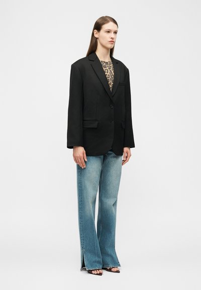 Žena s dlhými rovnými vlasmi, ktorá nosí nadmerne veľké čierne sako, top s leopardím vzorom, široké modré džínsy s bočnými rozparkami a čierne sandále s otvorenou špičkou.