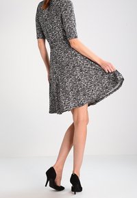 En svartvit mönstrad klänning med korta ärmar, figursydd midja och utsvängd kjol, kombinerad med svarta högklackade skor.