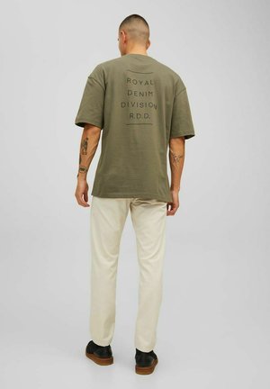 Camiseta oversized en color caqui con texto impreso en negro "ROYAL DENIM DIVISION R.D.D." en la parte de atrás, combinada con pantalones en color crema y zapatos negros.