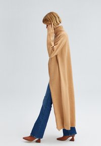 Lang, kamel-farvet rullekrave sweater lavet af blødt materiale, med en afslappet pasform og slidser i siderne, kombineret med flared denim og brune hælede støvler.