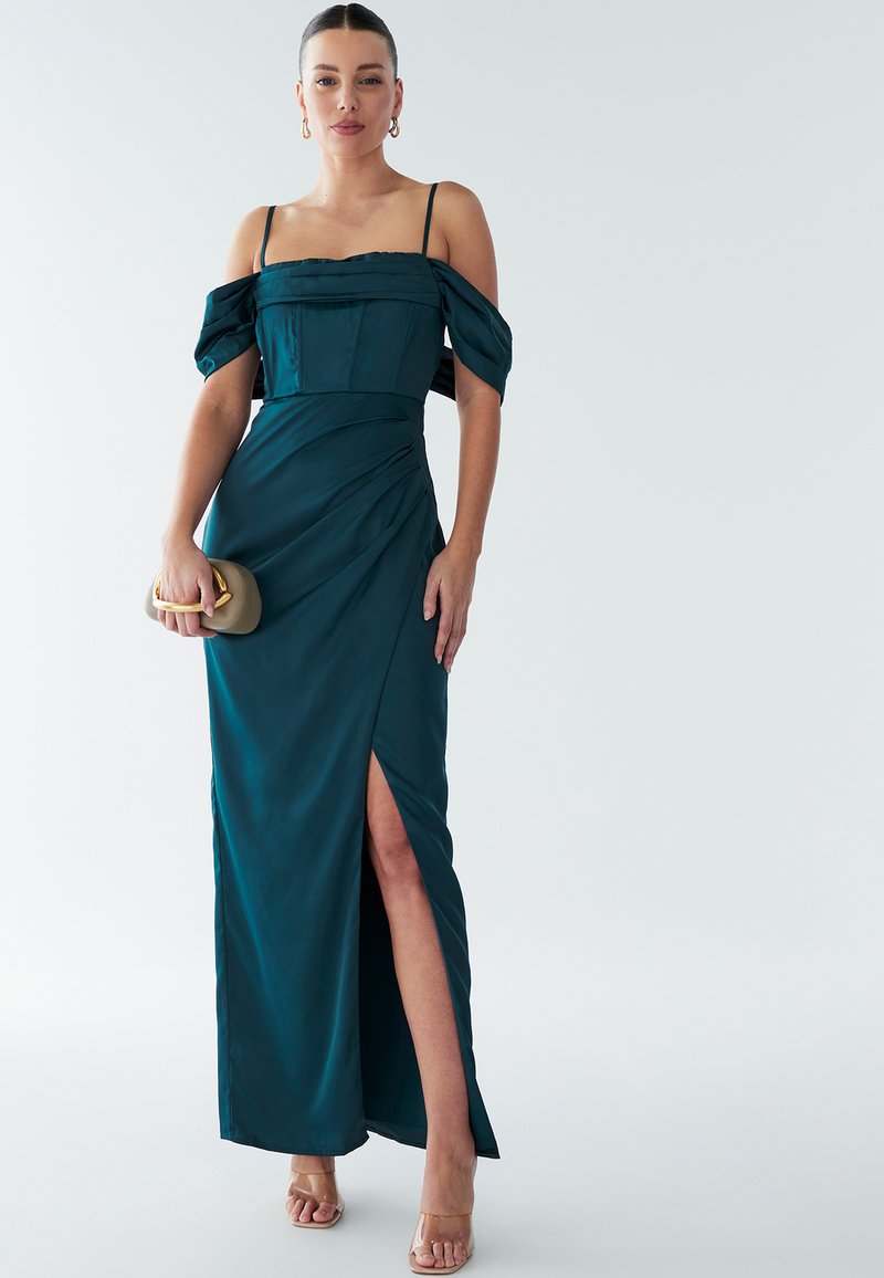 Teal satijnen jurk met off-the-shoulder pofmouwen, een getailleerde bodice en een zijsplit, gecombineerd met een gouden clutch en transparante hakken.