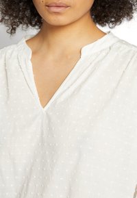 Blusa branca com decote em V, tecido texturizado com pequenos pontos bordados e pregas nos ombros para um ajuste descontraído.