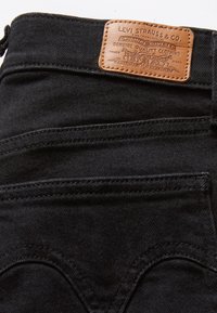 Poche arrière d'un jean en denim noir avec des détails cousus et un patch en cuir beige de la marque Levi Strauss & Co. au-dessus de la poche.