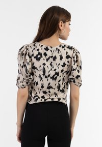 Beige Bluse mit schwarzem abstraktem Muster, gepufften kurzen Ärmeln, rundem Halsausschnitt und glattem Stoff. Getragen mit schmalen schwarzen Hosen.