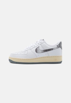 Hvid lavt snøresko med beige sål og grå tekstureret Nike-swoosh på siden og hælen mod en ensfarvet baggrund.
