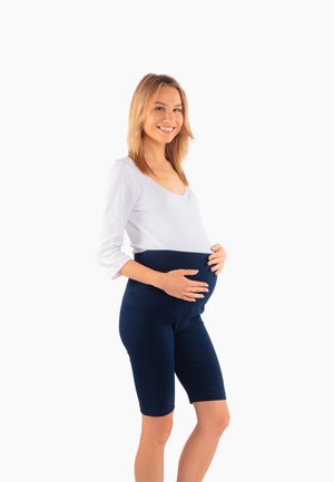 Uśmiechnięta kobieta w ciąży, ubrana w biały top i granatowe krótkie spodenki ciążowe, z delikatnie trzymającymi brzuch rękami.