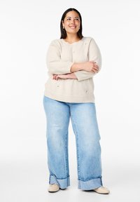 Beige Strickpullover mit floralem Stickdesign, kombiniert mit lockeren blauen Jeans aus Denim mit umgeschlagenem Saum. Die Schuhe sind hellfarbige Ballerinas.
