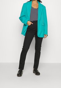 Donna che indossa un blazer turchese, una maglietta a righe bianche e nere, pantaloni neri e zoccoli neri con borchie, in piedi contro uno sfondo bianco.