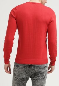 Pull en tricot rouge à manches longues, col rond et ourlet côtelé. Porté à l'envers, montrant une texture lisse et une coupe ajustée.