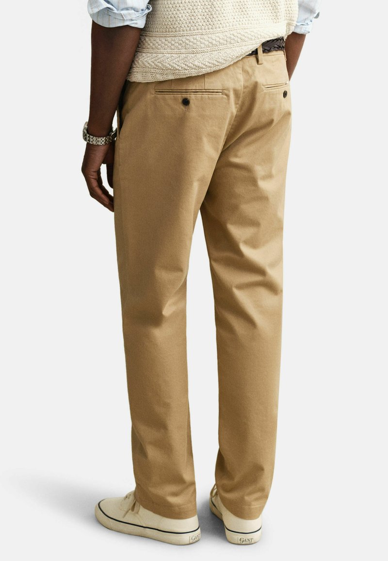 Hombre vistiendo pantalones chinos beige, zapatillas blancas, un chaleco de punto color crema y una camisa a rayas azul remangada, visto desde atrás.