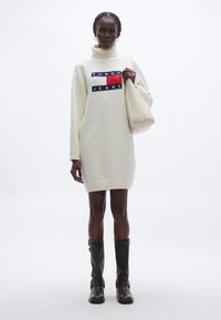 Krém színű kötött garbós ruha tengerészkék, piros és fehér logófolttal. Barna térdig érő csizmával és bézs színű tote táskával párosítva.