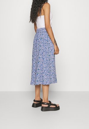 Vrouw draagt een wit tanktopje, een blauwe midi-rok met bloemenpatroon en zwarte plateauzandalen, staand zijwaarts tegen een effen lichte achtergrond.