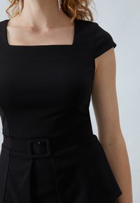 Robe noire ajustée avec des manches courtes, décolleté carré et taille ceinturée. Le tissu semble lisse avec un léger éclat, présentant des détails plissés.