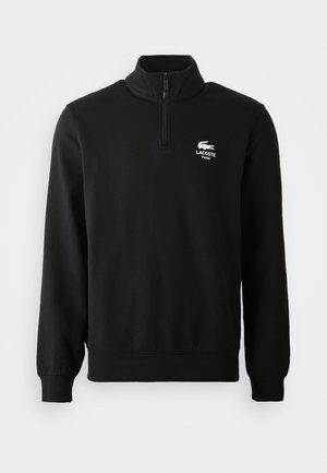 Sweatshirt noir Lacoste avec col haut et demi-fermeture éclair, comportant un logo blanc sur la poitrine gauche. Tissu doux avec des manches longues.