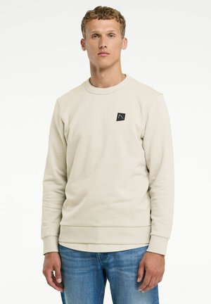 Jeune homme portant un sweat-shirt beige à col rond avec un petit logo noir sur la poitrine et un jean bleu, debout devant un fond blanc.