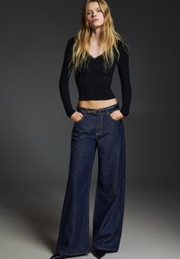 Crna majica s dugim rukavima uparena s tamnim trapericama širokih nogavica i visokog struka. Traperice imaju remen i dva prednja džepa, s diskretnim šavovima.