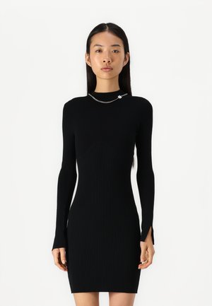 Zwarte ribbeljurk met lange mouwen, hoge hals en subtiele zijsplitten. Kenmerkt zich door een kleine zilveren kettingaccent bij de kraag.
