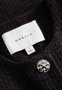 Λευκή ετικέτα ρούχου με "NOELLA" και μέγεθος "S Made in China" σε σκούρο υφασμάτινο ύφασμα με διακοσμητικό ασημένιο κουμπί κοντά.