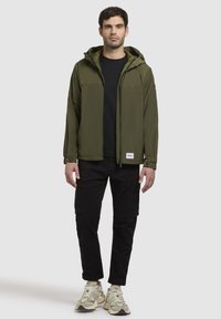 Herren olivgrüne Kapuzenjacke mit Reißverschlüssen, zwei Fronttaschen, kombiniert mit schwarzen Cargo-Hosen und hellen Sneakers. Glatte Stoffoberfläche.