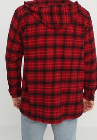Camicia in flanella a quadri rossi e neri con cappuccio, dotata di maniche lunghe e orlo curvo, realizzata in un tessuto testurizzato.